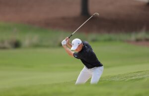 The Best Golf Books To Improve Your Game Golfer in a white cap and black shirt mid-swing on a green golf course fairway.