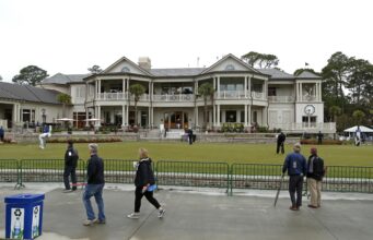 Does Club Golf Have a Pace of Play Problem? Golf practice area in front of a large clubhouse with balconies and palm trees, people walking and playing putt-putt on the green behind a green fence.