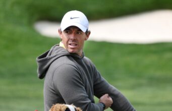 Rory McIlroy Reveals How He Got Devil Wears Prada 2 Role Man in a gray hoodie and white Nike cap looks to the side outdoors with a small plush dog visible in the foreground.