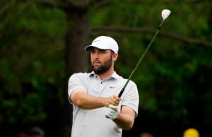 Scottie Scheffler Gives Sassy Response To LIV Golf Question Male golfer in a white cap and polo swinging a golf club on a grassy course, focused on the shot.