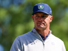 Bryson DeChambeau Just Admitted He Doesn’t Know What To Do Head-and-shoulders portrait of a man outdoors wearing a blue cap and a light gray zip-up sweater; patch on the cap shows a white skull and crossed bones, blurred green background.