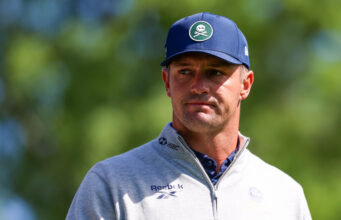 Bryson DeChambeau Just Admitted He Doesn’t Know What To Do Head-and-shoulders portrait of a man outdoors wearing a blue cap and a light gray zip-up sweater; patch on the cap shows a white skull and crossed bones, blurred green background.