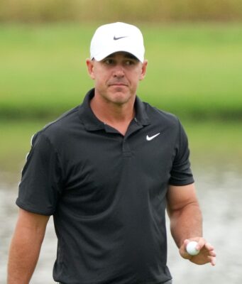 Koepka Drops Brutal Truth About LIV Golf’s Real Weakness Man in a black polo shirt and white Nike cap holding a golf ball on a golf course field.