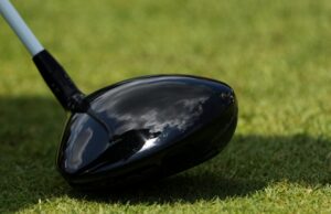Just Announced: Tour Edge Exotics Reveals New Mini Driver Tech Close-up of a glossy black golf driver head on green grass with a light gray shaft visible nearby.